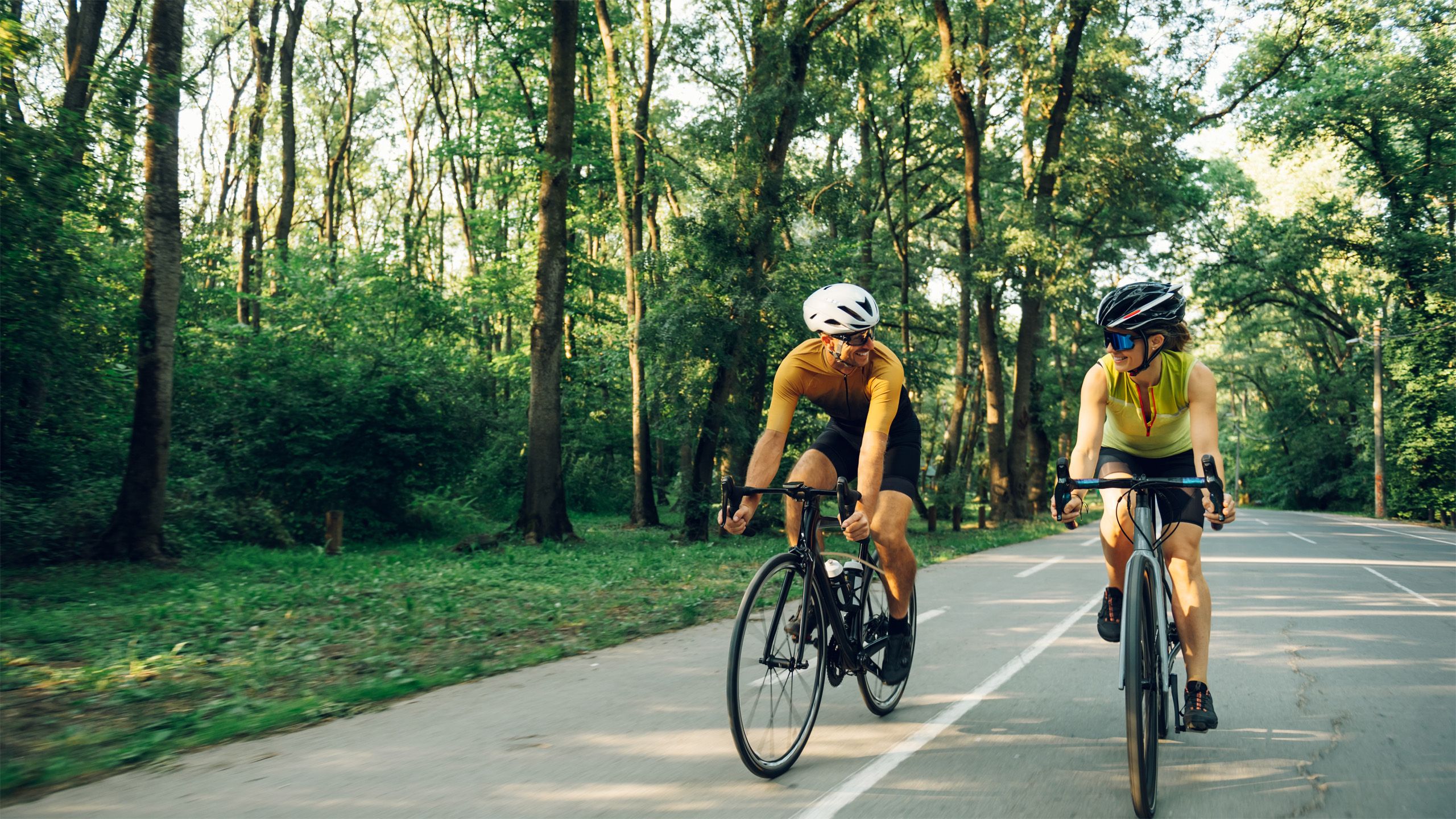 Zwei Radfahrer mit Helmen fahren auf einem asphaltierten Waldweg und unterhalten sich